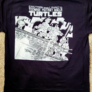 Könnte beinhalten: Marineblaues T-Shirt mit einer schwarz-weißen Grafik der Teenage Mutant Ninja Turtles. Der Text "Eastman and Laird's Teenage Mutant Ninja Turtles" ist oben auf dem Shirt aufgedruckt.
