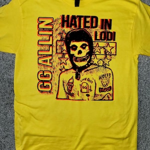 May include: Yellow t-shirt with a graphic design featuring a skull-like face and the text "HATED IN LODI" and "GG ALLIN" in red and black. The design includes stars and other graphic elements.