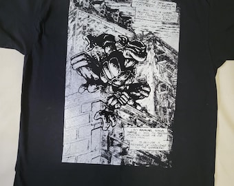 May include: Black and white graphic t-shirt featuring a Teenage Mutant Ninja Turtle character climbing a building. The shirt has a comic book style design with text that reads "I despise the damp, dank underground... but this is where I belong... second, my martial arts room to move about... I am Raphael, Ninja Turtle... and the Shredder chose me for this task... to deliver a message of challenge to the Shredder."