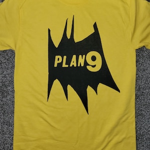 May include: Yellow t-shirt with a black graphic featuring the text "PLAN 9" in white. The graphic is a stylized, jagged shape. The shirt is made of a soft material.