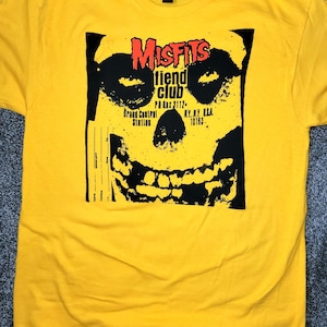 Op de afbeelding: Geel T-shirt met een zwart en rood grafisch ontwerp van een schedel en de tekst "Misfits Fiend Club". Het adres van Grand Central Station, NY, NY, is ook afgebeeld.