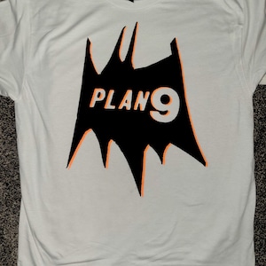 May include: A white t-shirt with a black graphic and orange detailing. The graphic is a jagged shape with the text "PLAN 9" in orange and white. The shirt has a crew neck and short sleeves.