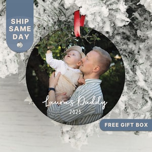 May include: Round Christmas ornament featuring a photo of a father and child. The ornament has a red ribbon for hanging. Text on the ornament reads "Laura's Daddy 2025". The background is a snowy tree.