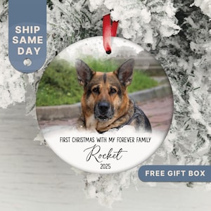 May include: A round, white Christmas ornament featuring a German Shepherd dog. The ornament has a red ribbon for hanging and includes the text "FIRST CHRISTMAS WITH MY FOREVER FAMILY" and the name "Rocket" with the year "2025".