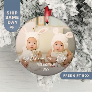 May include: Round Christmas ornament featuring two babies wearing bunny hats. The ornament has a photo of the babies and the text "Athena & Lily" and "First Christmas 2025". A red ribbon is attached for hanging.