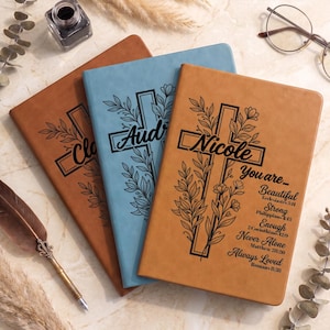 May include: Three rectangular journals in tan, light blue, and brown with smooth covers. Each features a black laser-engraved cross entwined with floral vines. The top journal displays the name Nicole, followed by the text You are... Beautiful, Strong, Enough, Never Alone, Always Loved, and Bible verse references.