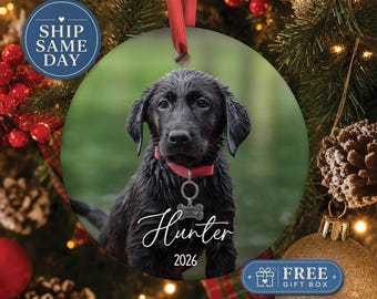 May include: A round ceramic ornament featuring a photograph of a black puppy wearing a red collar with a bone-shaped tag. The name Hunter is printed in white script below the puppy, followed by the year 2026. A red ribbon is attached to the top.