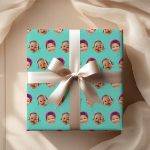 May include: Gift wrap featuring a repeating pattern of smiling baby faces wearing pink caps on a teal background. The wrapped gift is tied with a cream-colored satin ribbon and bow.