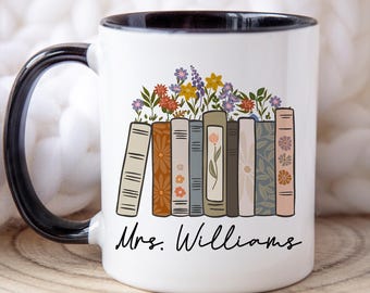 May include: White ceramic mug with a black handle and rim. The mug features a design of colorful flowers growing from a row of books. The name "Mrs. Williams" is written in black script below the books.