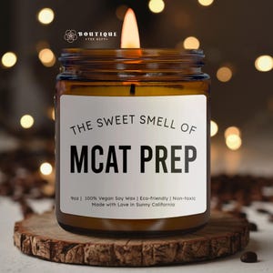 May include: A lit candle in a brown glass jar with a white label. The label reads "THE SWEET SMELL OF MCAT PREP." The candle is on a wooden coaster. The text on the label also says "9oz | 100% Vegan Soy Wax | Eco-friendly | Non-toxic Made with Love in Sunny California."