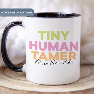 May include: White ceramic coffee mug with a black rim and handle. The mug has the text "TINY HUMAN TAMER Ms. Smith" in colorful letters.