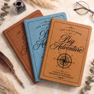 May include: Three rectangular journals in tan, light blue, and brown with black text reading THE LITTLE BOOK of Big Adventure and A SPACE TO RECORD MEMORIES OF WANDERLUST, featuring a compass rose graphic on the front covers.