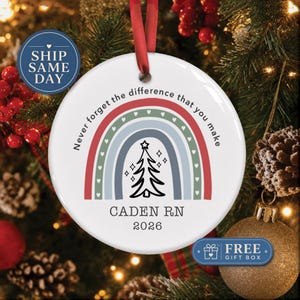 May include: A round white ceramic Christmas ornament featuring a colorful rainbow graphic with small hearts, a black line-art Christmas tree, and the text "Never forget the difference that you make", "CADEN RN", and "2026" printed in black.