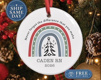 May include: A round white ceramic Christmas ornament featuring a colorful rainbow graphic with small hearts, a black line-art Christmas tree, and the text "Never forget the difference that you make", "CADEN RN", and "2026" printed in black.