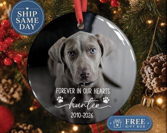 May include: A round, glossy ceramic ornament featuring a photograph of a gray puppy with blue eyes. The text reads FOREVER IN OUR HEARTS, hunter, and 2010-2026, accompanied by two paw print icons. A red ribbon is attached to the top for hanging.
