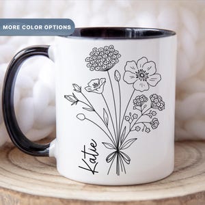 May include: White ceramic mug with a black rim and handle. The mug features a black line drawing of a bouquet of flowers and the name "Katie" at the bottom.