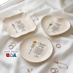May include: Three cream-colored ceramic trinket dishes with gold-trimmed edges, featuring floral illustrations and the text Chapter Sixty, Est. 1966, and personalized names. The dishes are square, heart-shaped, and round, displayed with jewelry.