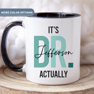May include: White ceramic coffee mug with a black rim and handle. The mug has a teal green design that says "It's Dr. Jefferson Actually".