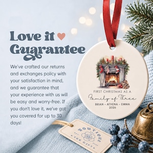 May include: A round white ceramic ornament with a red ribbon, featuring a fireplace illustration, the text "FIRST CHRISTMAS AS A FAMILY OF THREE", the names "BRIAN - ATHENA - EMMA", and the year "2026". A small beige tag with "BOUTIQUE TEE GIFT" is nearby.