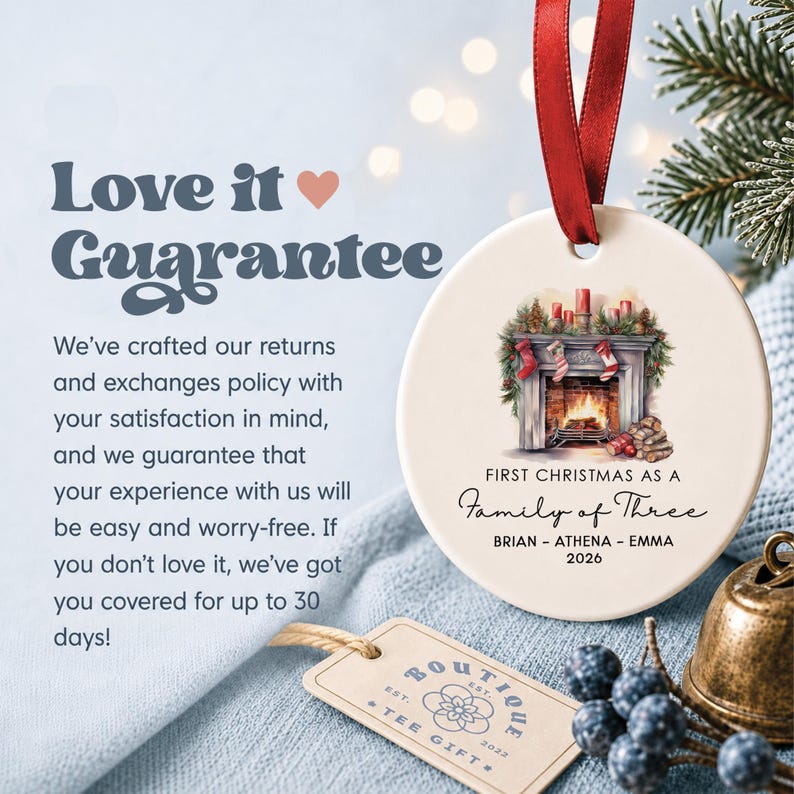 May include: A round white ceramic ornament with a red ribbon, featuring a fireplace illustration, the text "FIRST CHRISTMAS AS A FAMILY OF THREE", the names "BRIAN - ATHENA - EMMA", and the year "2026". A small beige tag with "BOUTIQUE TEE GIFT" is nearby.