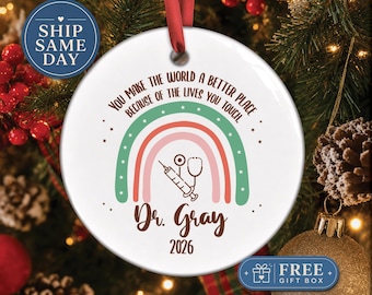 Puede incluir: Un adorno redondo de cerámica blanca con una cinta roja. El diseño presenta un arcoíris en tonos pastel, una jeringa y un estetoscopio. El texto dice: You make the world a better place because of the lives you touch. Dr. Gray 2026.