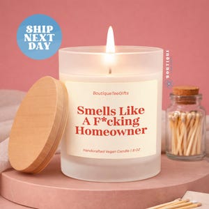 May include: A lit, frosted glass candle with a wooden lid. The candle has a label that reads "Smells Like A F*cking Homeowner." A small jar of matches sits nearby. The candle is 8 oz.