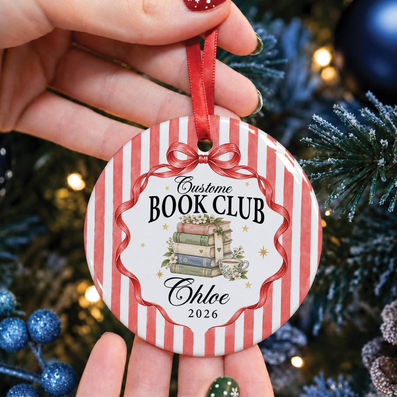 May include: A round ceramic Christmas ornament featuring a red and white striped background. The center displays a stack of books, a teacup, and the text "Custome BOOK CLUB", "Chloe", and "2026". A red ribbon loop is attached to the top for hanging.