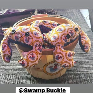 May include: A light-colored basket filled with several plush toys shaped like crabs. The crab toys are made of fabric with a repeating pattern of a rooster inside a circle. The toys have button eyes and are labeled "Swamp Buckle".