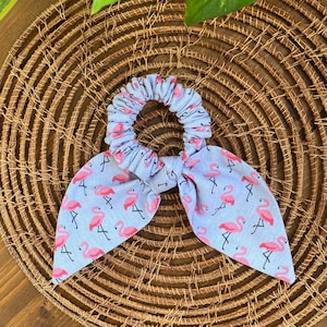 May include: A light blue scrunchie with a pink flamingo pattern and a matching bow. The scrunchie is made of fabric and is designed to hold hair. The background is a woven basket.