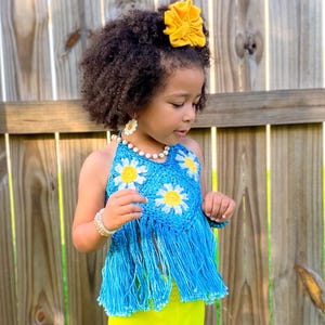 May include: A young girl wearing a blue crochet top with daisy accents and fringe, paired with bright yellow bell-bottom pants. A yellow flower hair accessory completes the outfit. The ensemble is set against a wooden fence and green grass.