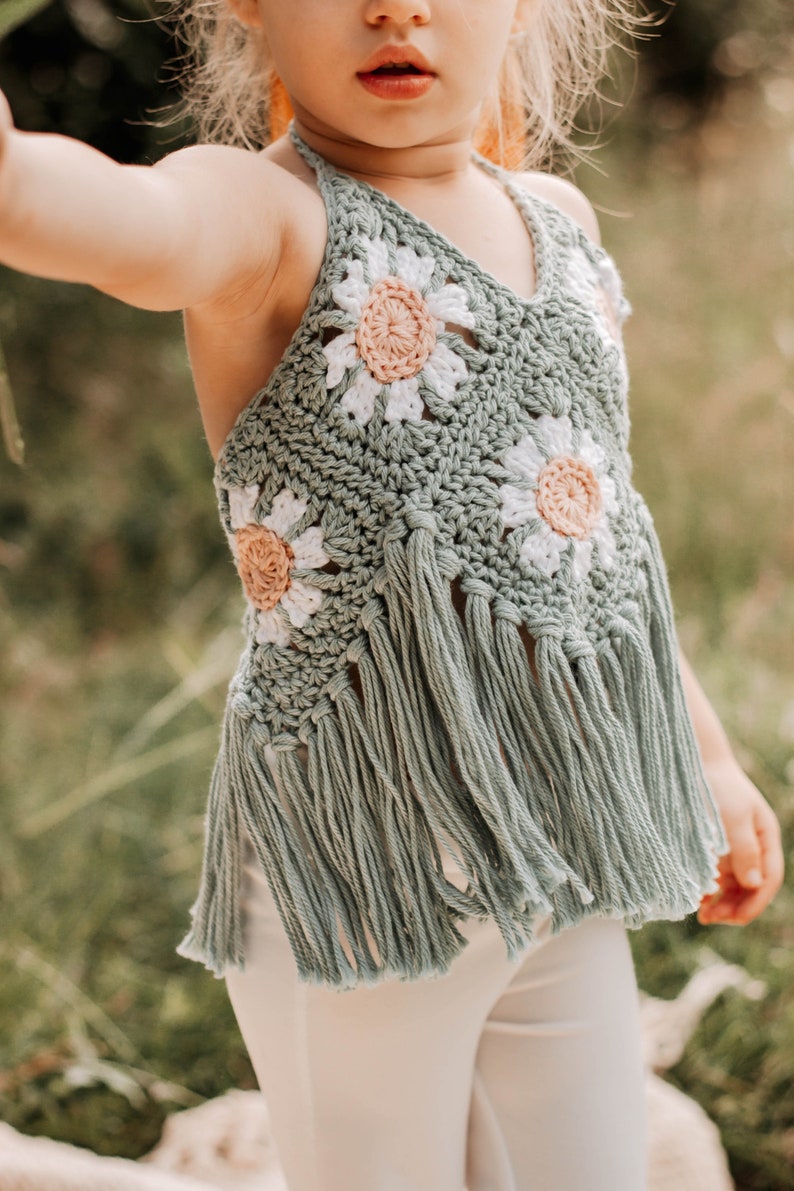 K&ouml;nnte beinhalten: Ein hellgr&uuml;nes H&auml;kel-Halter-Top mit wei&szlig;en und rosa G&auml;nsebl&uuml;mchen und Fransen. Das Top wird von einem Kind getragen.