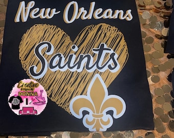 Puede incluir: Camiseta negra con las palabras "New Orleans Saints" en blanco y dorado. También se muestra un corazón dorado con líneas garabateadas y una flor de lis. La camiseta tiene un logotipo en la esquina inferior izquierda.