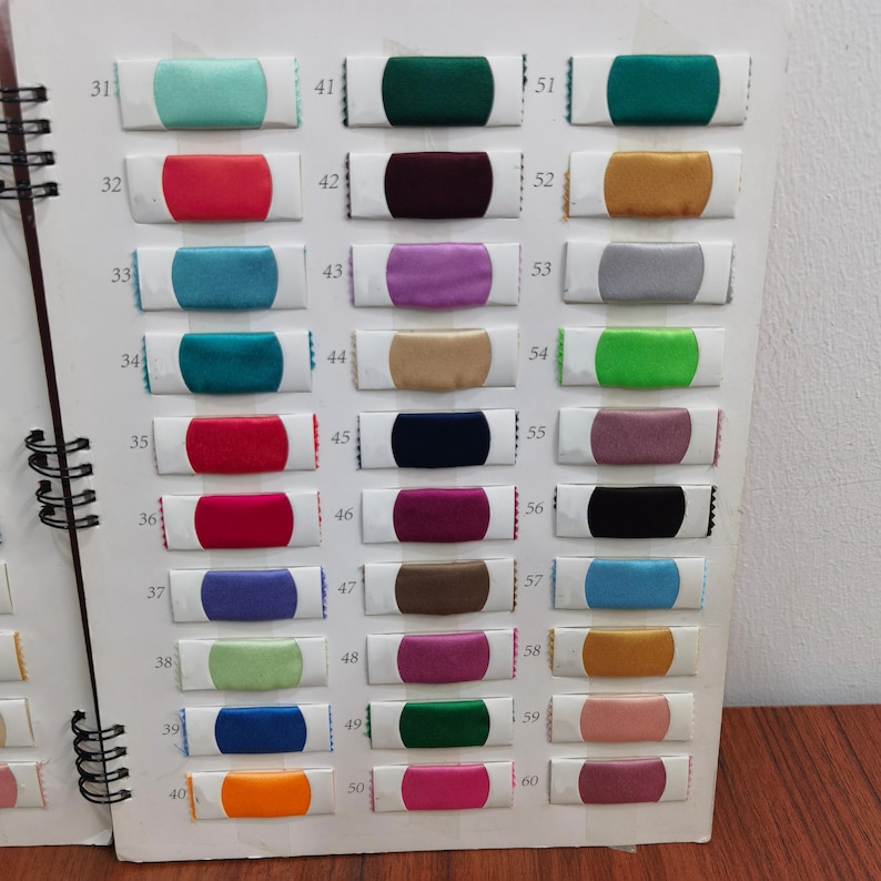 K&ouml;nnte beinhalten: Ein Farbmusterbuch mit Spiralbindung, das zahlreiche Stoffmuster in verschiedenen Farben zeigt. Die rechteckigen Muster weisen Farbt&ouml;ne wie T&uuml;rkis, Rot, Lila und Gold auf. Das Buch ist ge&ouml;ffnet und zeigt mehrere Reihen von Farboptionen.