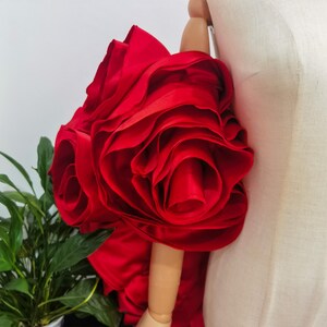 K&ouml;nnte beinhalten: Eine gro&szlig;e, leuchtend rote Stoffrose, m&ouml;glicherweise ein dekoratives Element f&uuml;r Kleidung oder Accessoires. Die Rose besteht aus geschichtetem, ger&uuml;schtem Material, wodurch ein voller, strukturierter Eindruck entsteht. Die Bl&uuml;tenbl&auml;tter sind tiefrot.