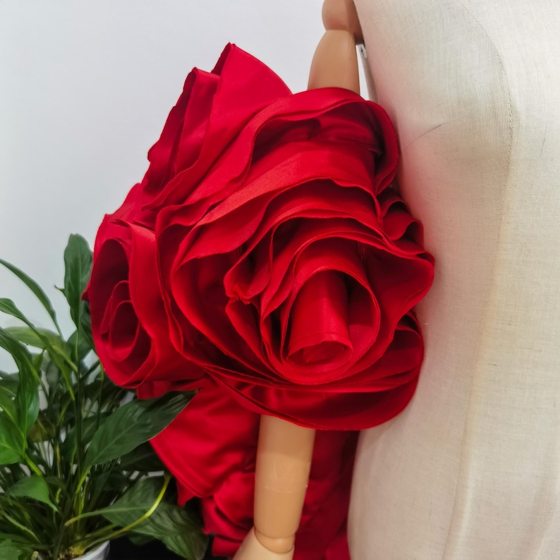K&ouml;nnte beinhalten: Eine gro&szlig;e, leuchtend rote Stoffrose, m&ouml;glicherweise ein dekoratives Element f&uuml;r Kleidung oder Accessoires. Die Rose besteht aus geschichtetem, ger&uuml;schtem Material, wodurch ein voller, strukturierter Eindruck entsteht. Die Bl&uuml;tenbl&auml;tter sind tiefrot.