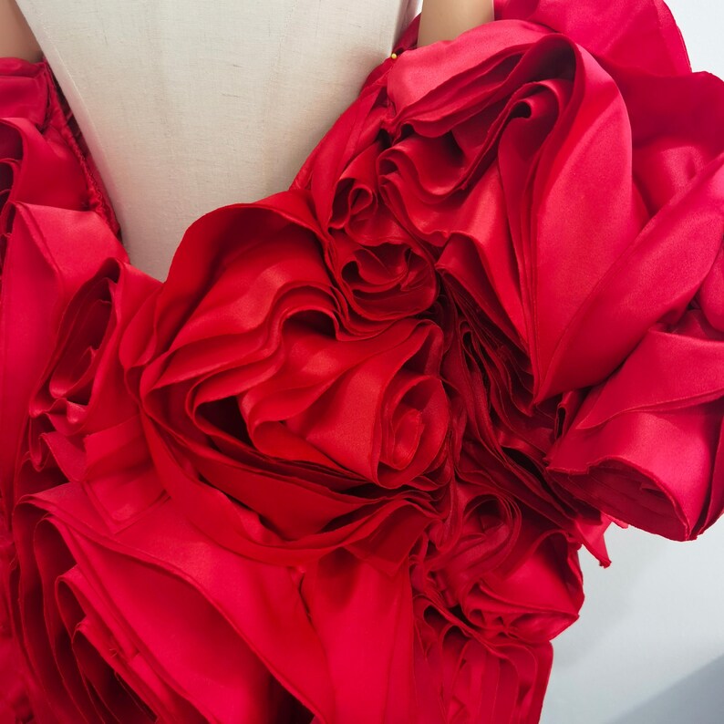 K&ouml;nnte beinhalten: Ein leuchtend rotes Satinkleid mit aufwendigen, geschichteten floralen Details. Der Stoff ist gerafft und geformt, um gro&szlig;e, dimensionale Rosetten zu erzeugen. Das Kleid wird auf einer Schaufensterpuppe pr&auml;sentiert, die die dramatische, flie&szlig;ende Silhouette hervorhebt.