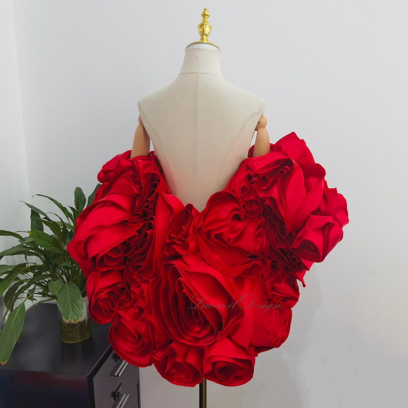 K&ouml;nnte beinhalten: Ein leuchtend rotes, ger&uuml;schtes Kleidungsst&uuml;ck, m&ouml;glicherweise ein Oberteil oder ein Kleidungsdetail, wird auf einer Schaufensterpuppe pr&auml;sentiert. Der Stoff ist gerafft und geschichtet, wodurch ein volumin&ouml;ser, bl&uuml;tenartiger Effekt entsteht. Die Schaufensterpuppe hat einen goldfarbenen Kopf und Holz&auml;rmel.