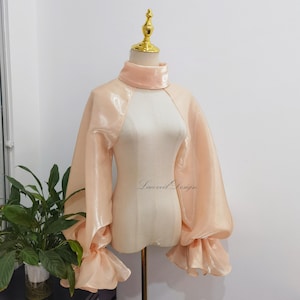 May include: A peach-colored, sheer bolero top with a high collar and voluminous sleeves. The sleeves gather at the wrists into ruffled cuffs. The top is displayed on a mannequin with a gold-colored finial. The text "Lacevil Design" is visible.