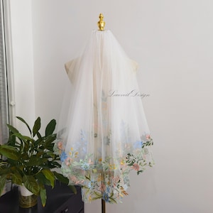 Peut inclure: Un voile de mariée blanc avec une bordure brodée de fleurs aux couleurs pastel. Le voile est présenté sur un mannequin, mettant en valeur le design délicat et le texte "Laceveil Design".