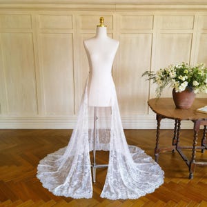 May include: A white lace bridal skirt with a long train is displayed on a dress form. The skirt features a delicate floral pattern and a scalloped edge. The dress form is on a metal stand, and the background includes a wooden floor and wall.