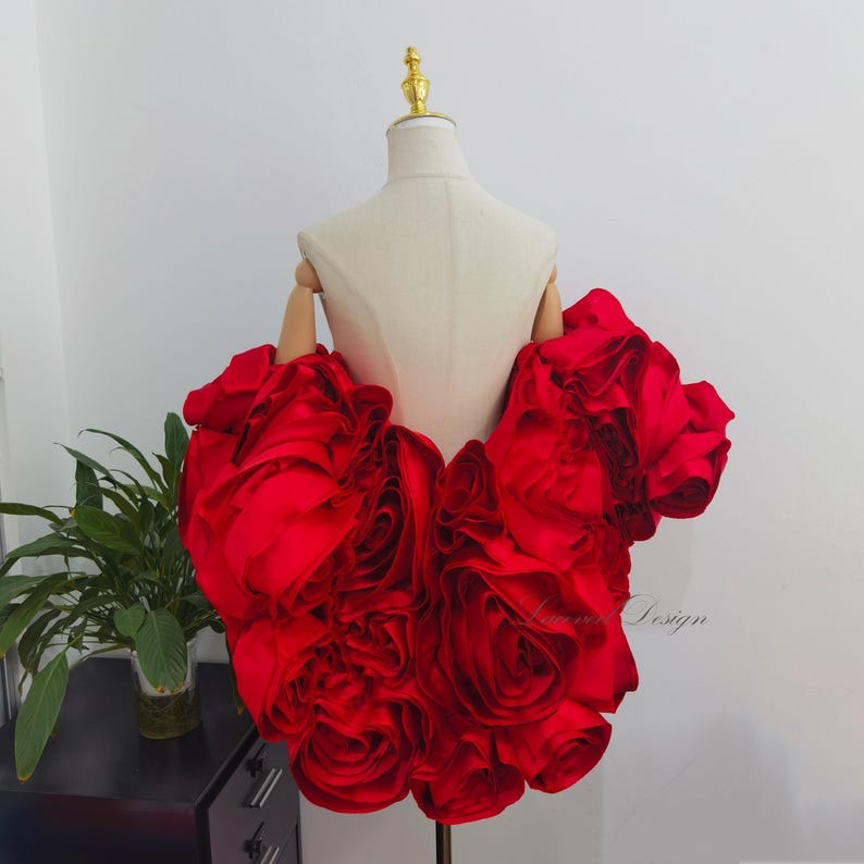 K&ouml;nnte beinhalten: Ein leuchtend rotes, rosenf&ouml;rmiges Kleidungsst&uuml;ck, wahrscheinlich ein Oberteil oder ein Kleidungsdetail, wird auf einer Schaufensterpuppe pr&auml;sentiert. Der Stoff ist gerafft und geschichtet, um einen volumin&ouml;sen, floralen Effekt zu erzeugen. Die Schaufensterpuppe hat einen goldenen Aufsatz und Holz&auml;rmel.