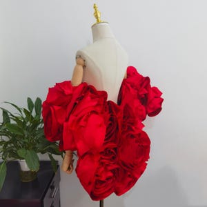 K&ouml;nnte beinhalten: Ein rotes, mit Rosen geschm&uuml;cktes Kleidungsst&uuml;ck, das auf einer Schaufensterpuppe pr&auml;sentiert wird. Das Design des Kleidungsst&uuml;cks zeigt gro&szlig;e, ger&uuml;schte Stoffrosen, die ein volumin&ouml;ses und strukturiertes Aussehen erzeugen. Die Schaufensterpuppe hat einen cremefarbenen Torso und Holz&auml;rmel.
