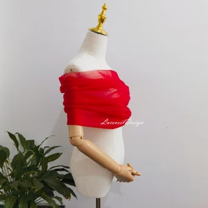 Puede incluir: Un pañuelo de tela rojo vibrante y semitransparente drapeado horizontalmente sobre el pecho de un maniquí blanco. El material ligero presenta pliegues suaves y una textura delicada. El texto Laceveil Design está impreso en blanco.
