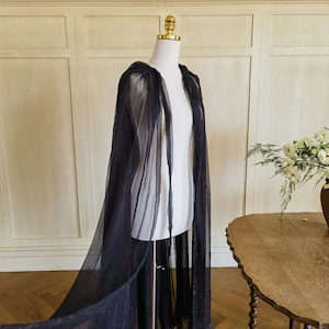 Puede incluir: Una capa transparente de color azul marino oscuro con capucha, drapeada sobre un maniquí blanco. La capa está hecha de una tela ligera y fluida. La capa se exhibe sobre un soporte metálico. El fondo incluye una pared de madera y una mesa con un jarrón de flores.