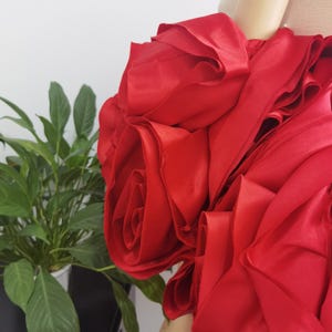 K&ouml;nnte beinhalten: Ein leuchtend rotes Stoffkleid mit gro&szlig;en, rosenartigen Petalldetails. Das Kleid hat ein mehrschichtiges Design, das ein volumin&ouml;ses und strukturiertes Aussehen erzeugt. Die leuchtend rote Farbe ist auff&auml;llig, und der Stoff hat einen leichten Glanz.