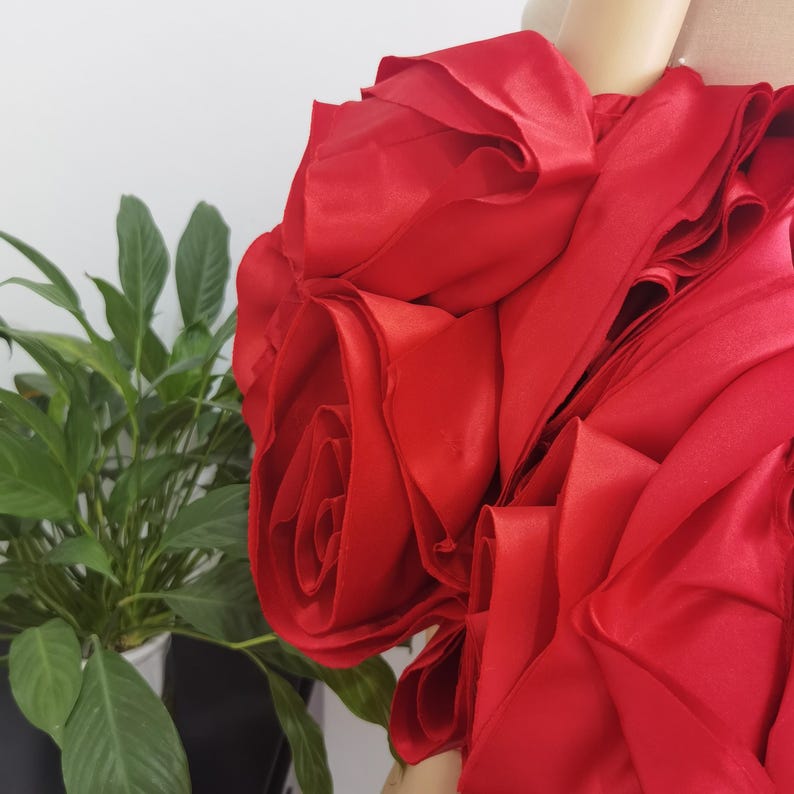 K&ouml;nnte beinhalten: Ein leuchtend rotes Stoffkleid mit gro&szlig;en, rosenartigen Petalldetails. Das Kleid hat ein mehrschichtiges Design, das ein volumin&ouml;ses und strukturiertes Aussehen erzeugt. Die leuchtend rote Farbe ist auff&auml;llig, und der Stoff hat einen leichten Glanz.