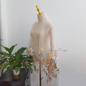 Peut inclure: Un voile de mariée ivoire transparent avec broderies florales aux couleurs pastel. Le voile est drapé sur un mannequin, mettant en valeur le tissu délicat et les détails floraux complexes. Le texte "Laceveil Design" est visible sur le voile.