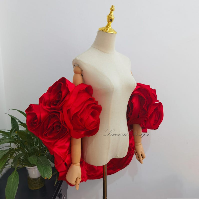 K&ouml;nnte beinhalten: Eine Schaufensterpuppe pr&auml;sentiert ein dramatisches rotes Satinkleid mit gro&szlig;en, rosenf&ouml;rmigen Verzierungen an den &Auml;rmeln. Das Design hat einen High-Low-Saum und ist vor einem neutralen Hintergrund platziert. Der Text "Laceveil Design" ist sichtbar.