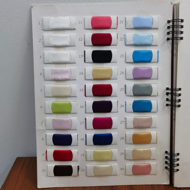 K&ouml;nnte beinhalten: Ein Farbmusterbuch mit 30 Stoffmustern in verschiedenen Farben. Die Muster sind in Reihen und Spalten angeordnet, wobei jedes Muster mit einer Nummer versehen ist. Zu den Farben geh&ouml;ren Wei&szlig;, Rosa, Rot, Blau und Gold.