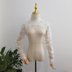 Peut inclure: Un boléro en dentelle blanche avec un col montant et des manches longues ornées de broderies florales complexes. Le vêtement présente de petites perles sur les poignets et est présenté sur un mannequin avec un embout doré au cou.